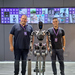 Zwei junge Männer stehen rechts und links von einem humanoiden Roboter, im Hintergrund sind Bildschirme zu sehen