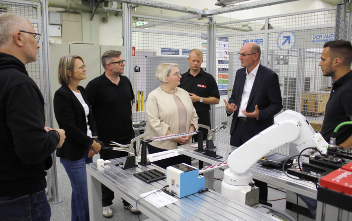 Eine Gruppe von Menschen steht um einen Tisch mit einerm Roboterarm herum, ein Mann erklärt etwas 